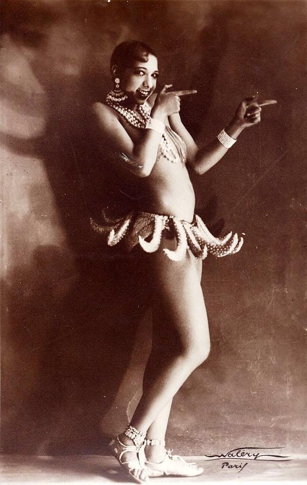Image is a sepia-tone photo of Josephine Baker, a young black woman. She's wearing several necklaces that appear to be strings of huge pearls, a sort of tutu made from spotted bananas, and huge fan-shaped earrings. She's standing in profile pointing to the right with her index fingers.