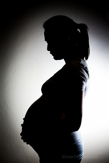 Image shows a woman in shadow, holding her pregnant belly. There is a circle of light on the white wall behind her.