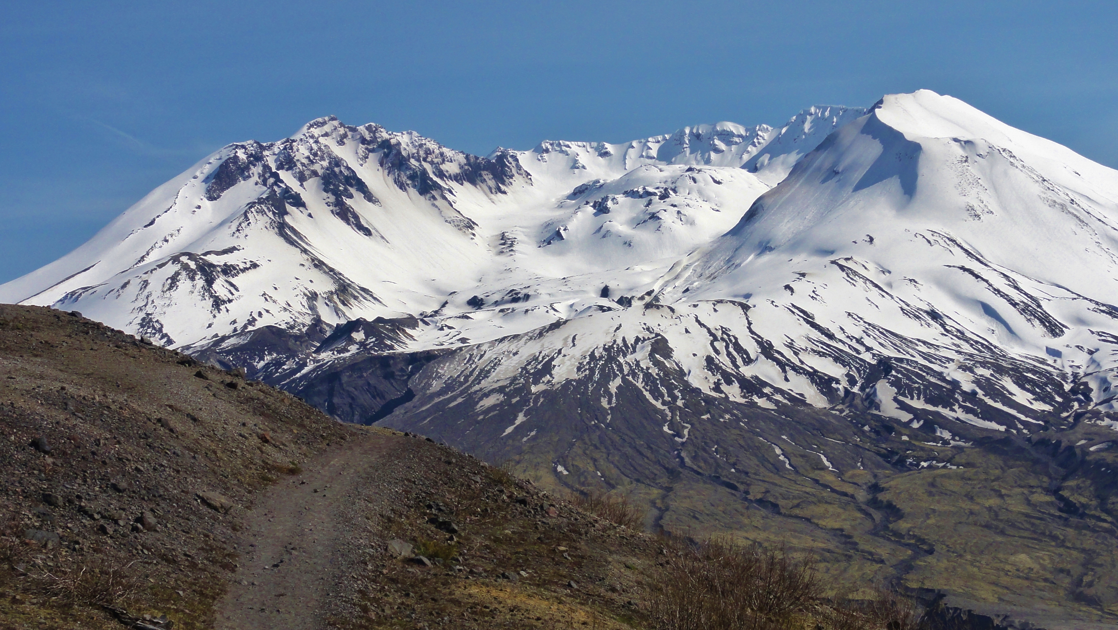Image shows a bit of dirt trail winding along the side of a bare ridge. It seems to vanish into the snow-capped crater of Mount St. Helens in the distance. The view shows the dome rising from the center.