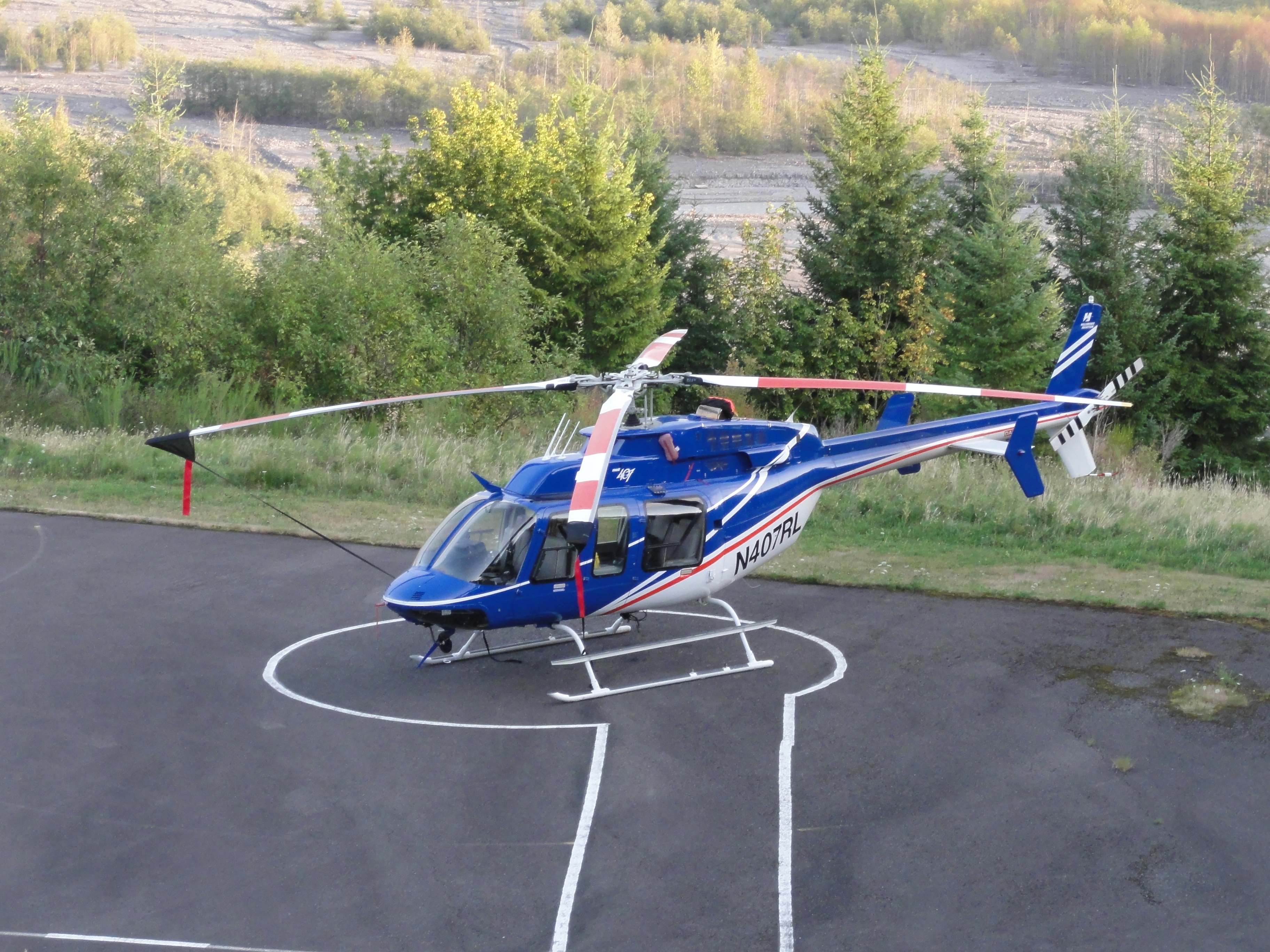 Image shows a blue helicopter with a white underbelly parked on a helipad. The Toutle River valley is visible behind it.