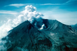 Image shows the remains of Mount St. Helens's summit. The volcano is covered in nearly-black ash. There is a notch in the crater wall where the lateral blast occurred. Steam rises from the vent and spills over the south rim, which is toward the back left of the photo. 