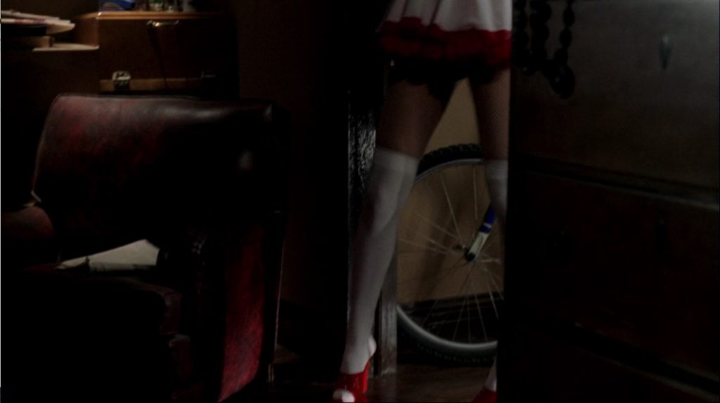 Image shows a woman's legs stepping from one room to the next. She's wearing red high heels over knee-high white stockings. The edge of a short, flaring white dress with red trim is visible. To the right, part of a chest of drawers can be seen. Behind her, the tire of a bike is visible. To the left, a maroon easy chair can be seen. The whole scene is dimly lit.