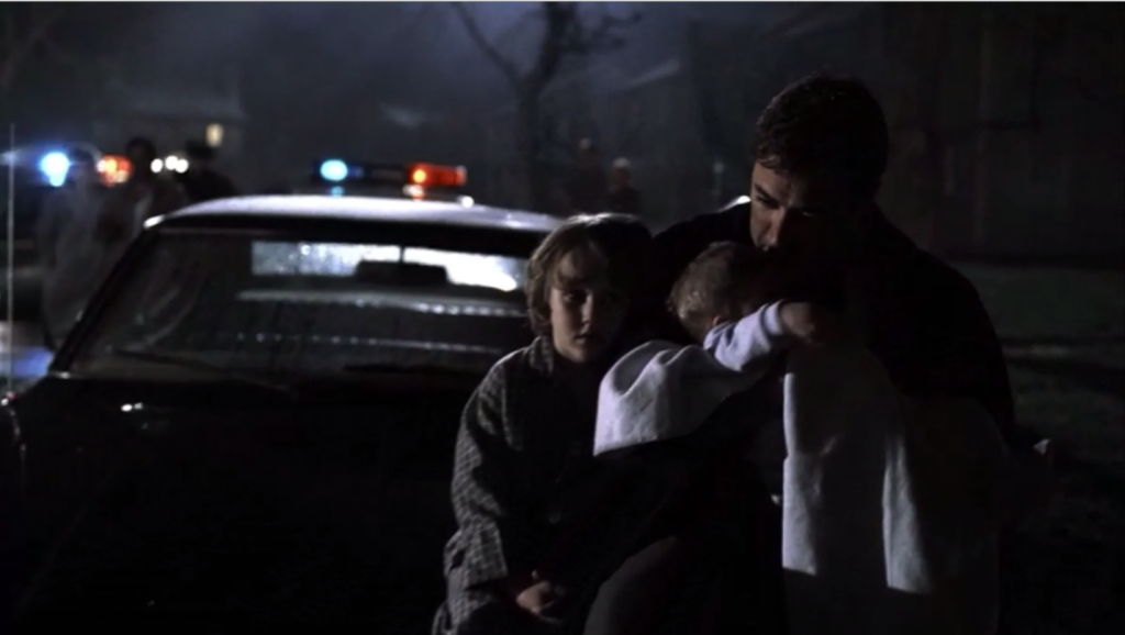 Image shows the Winchester family sitting on the hood of a police car. Dean is leaning against his dad, who holds baby Sam.