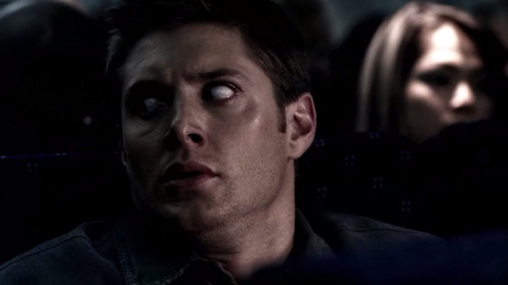 Screenshot shows Dean in an airplane seat, head turned toward the left, looking at something off-camera. He's so scared almost all we can see are the whites of his eyes. His face is frozen in an expression of dread.