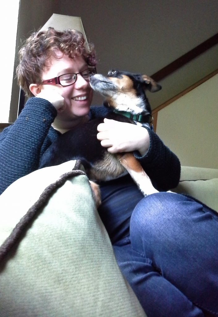 Image shows Pipa, a minature pinscher, standing in Aoife's lap and giving her a kiss on the nose while Aoife grins.