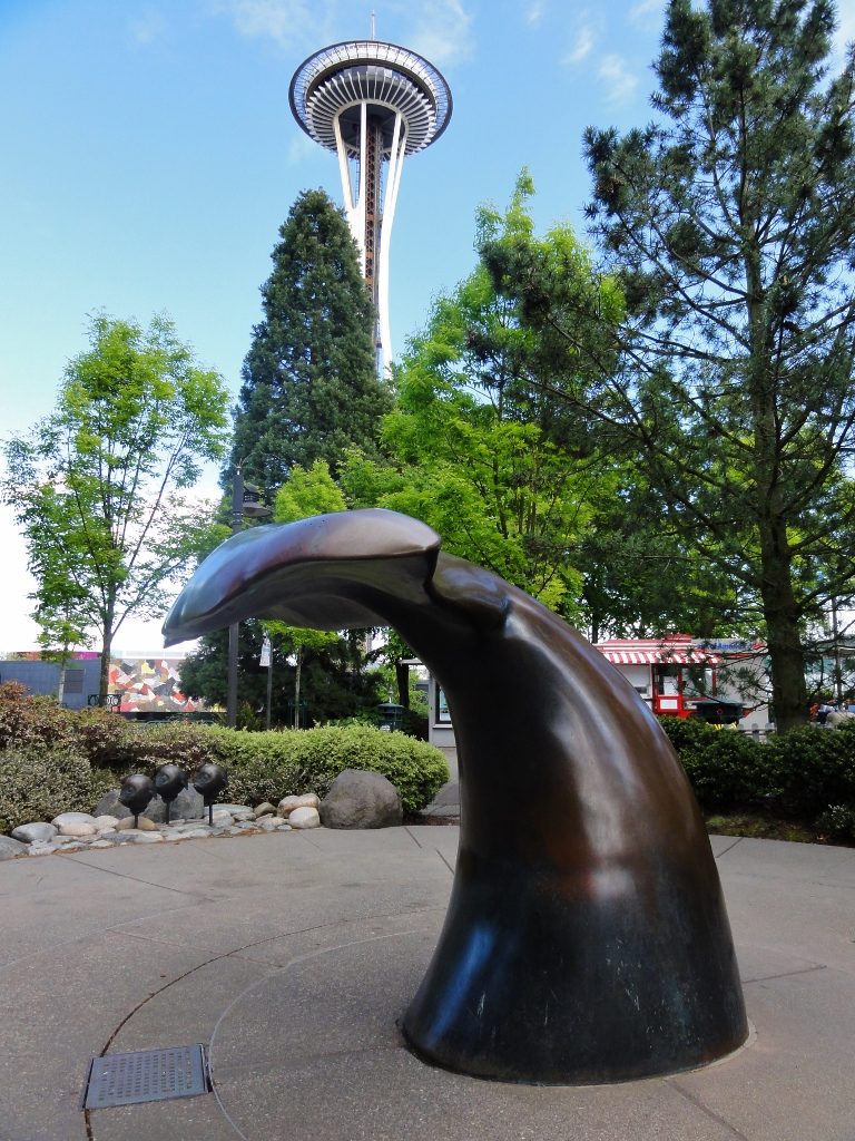 Image shows a whale's tail, curved as if the animal is diving, with the Space Needle in the background just past a screen of various trees.