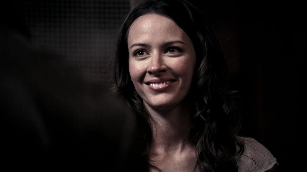 Screenshot from the episode shows Amy Acer, a brunette woman, from the neck up, smiling. She's surrounded by darkness.