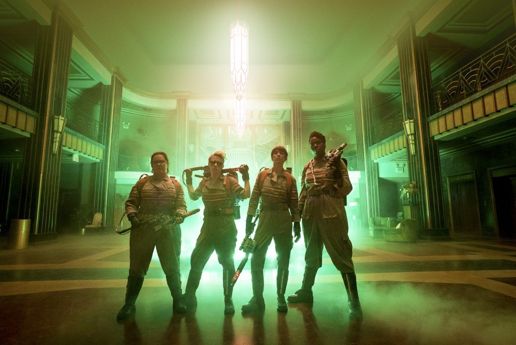 Promotional photo for the new Ghostbusters, showing the team standing with their equipment. They're in a large open area in a building, with a glowing green light source behind them.