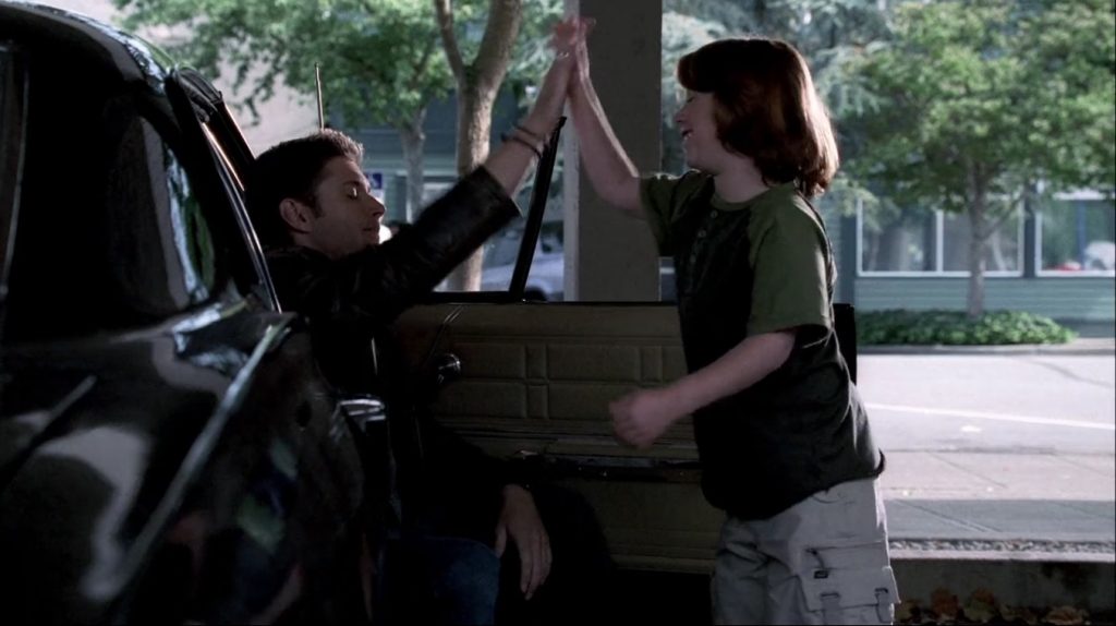 Image shows Dean sitting sideways on the passenger seat of the Impala with the door open. Lucas is standing in front of him. They're sharing an enthusiastic high five.