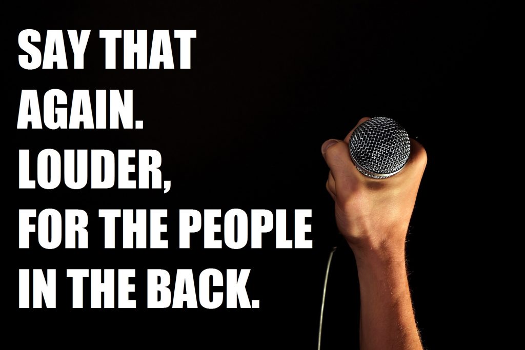 Image shows a white hand holding a microphone towards the camera. It's in the bottom right of a black background. Caption in white letters beside it says, "Say that again. Louder, for the people in the back."