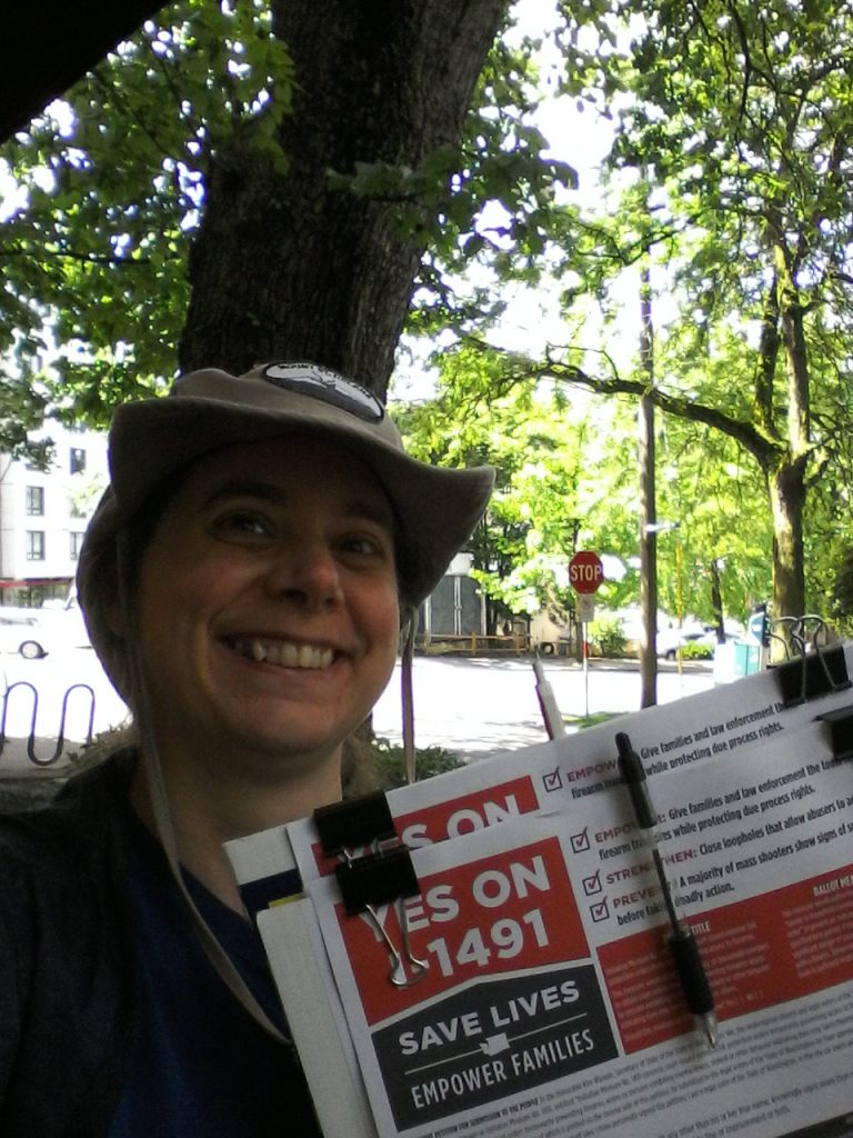 Image shows me standing by a tree, with a street and buildings visible in the sunshine behind me. I've got my tan Mount St. Helens hat on. I'm holding petitions that say "Yes on I-1491. Save Lives. Empower Families."