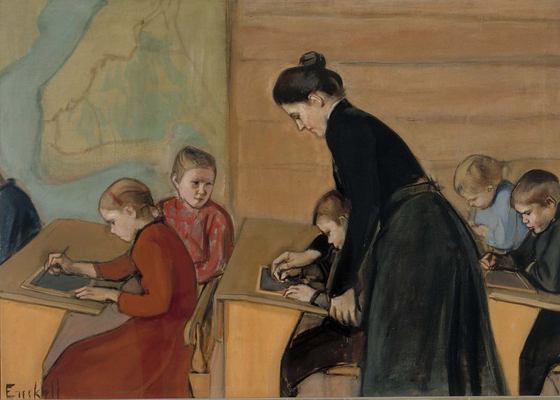 Image is a painting showing a school room. There is a map on the wall. Young children are sitting in long wooden desks. The girls have on long dresses, most of them red or light blue. The boys have on black suits. The teacher is leaning over helping one of the boys. She is dressed in a black blouse and long black skirt with her dark hair up in a bun.