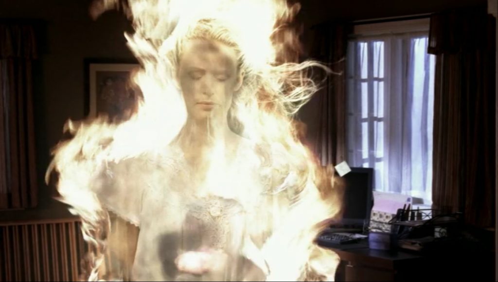 Image shows Mary Winchester, a blonde woman in a white nightgown. She is looking down with her eyes closed. She is engulfed in ghostly flames.