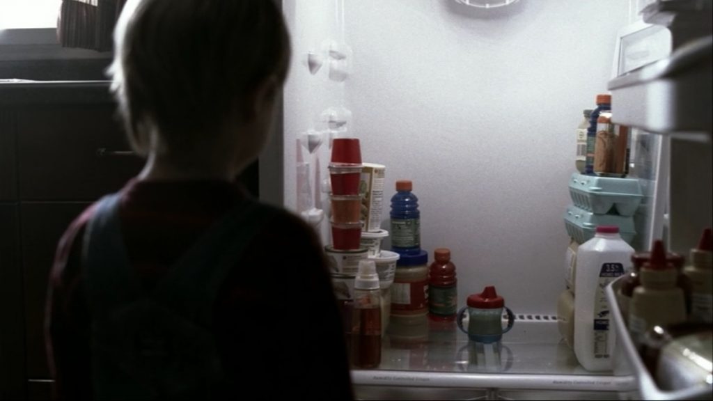 Image is looking into a fridge. Stuff is stacked up to either side of a two-handled sippy-cup full of juice. We can see a tow-headed toddler standing in the open door with his back to us, staring at the juice.