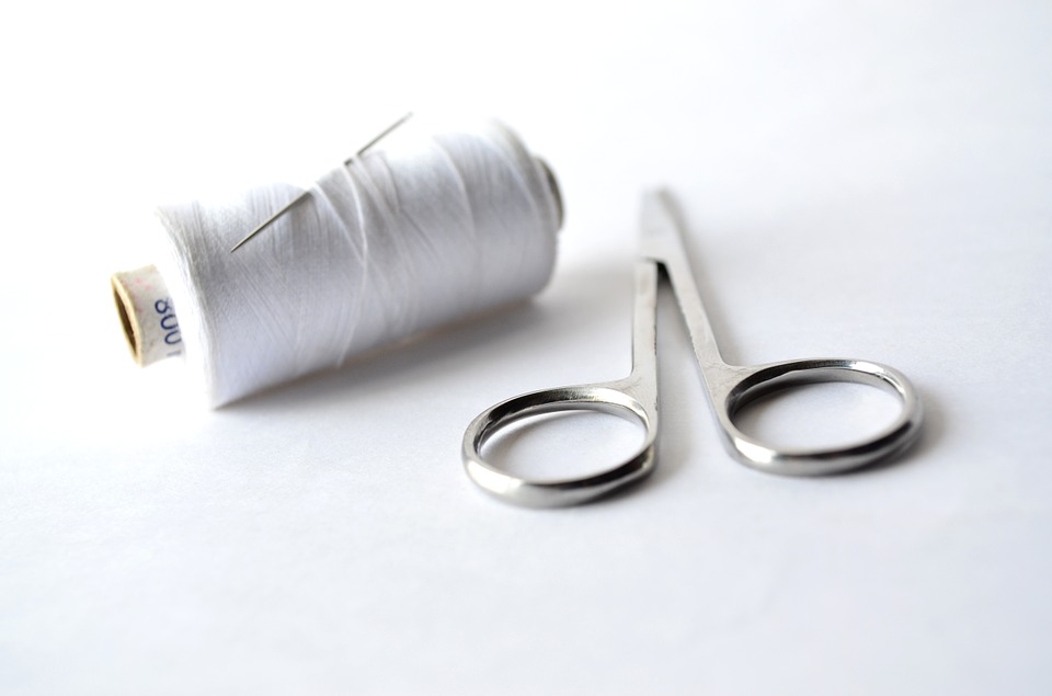 Image is a roll of white sewing thread that looks a bit like floss, with a straight sewing needle stuck through it. Beside it is a pair of silver-handled small sewing and craft scissors.