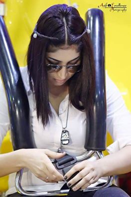 Image shows Qandeel Baloch, a young woman with long dark hair, wearing sunglasses and looking down. She is buckling herself into what looks like a carnival ride. There are black bars over her shoulders like one sees on roller coasters.