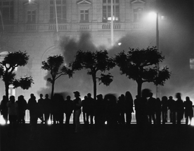 Black and white image shows a ghostly building with high windows and balconies in the background. There is a line of skinny trees whose trunks end in a puff of branches. Below the trees is a line of silhouetted figures standing in a line. The scene is lit by a streetlamp whose light seems to be diffused by smoke or mist.