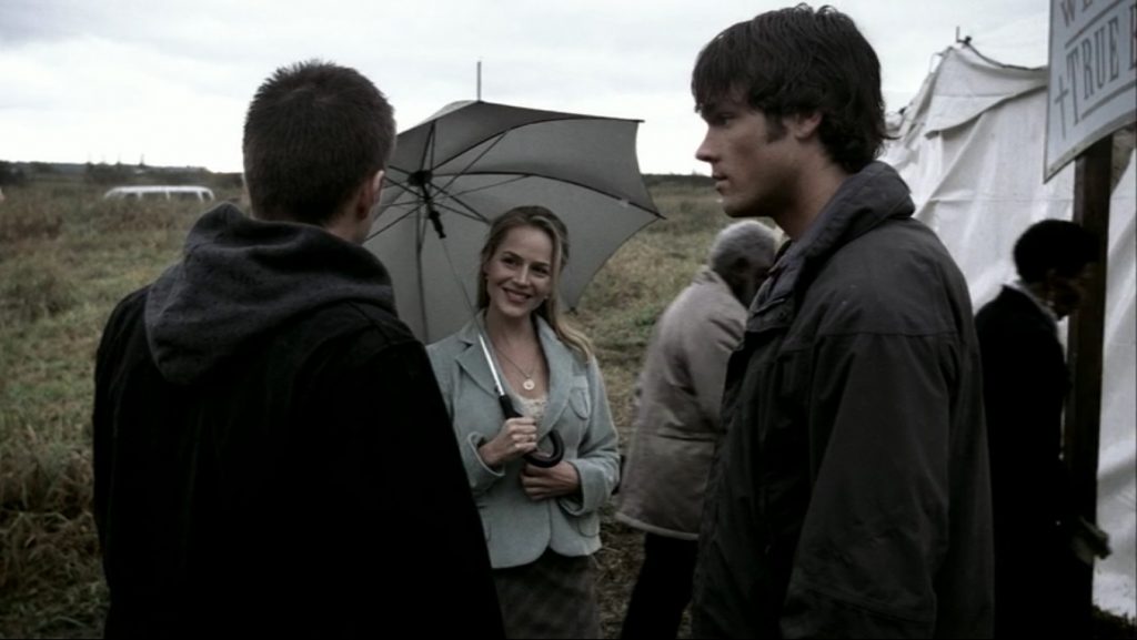 Image shows a blonde woman with a charming smile holding an umbrella and looking at Dean, who has his back to the camera. Sam is in profile beside him.