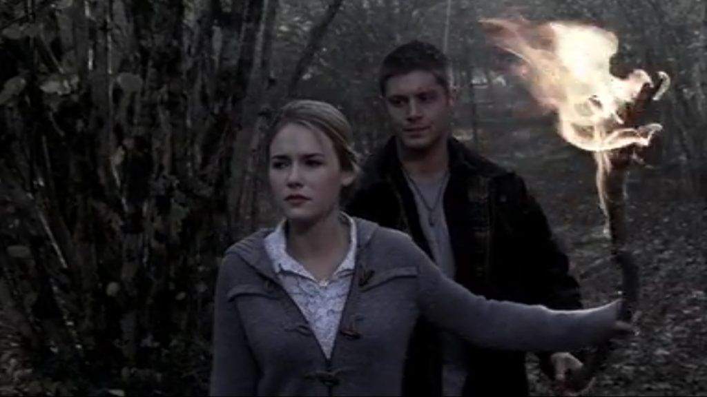 Image shows Emily, a young woman with long, blond hair pulled back in a pony tail. She's holding a flaming branch in one extended hand and is looking grimly determined. Dean is over her shoulder with a you-do-you expression.