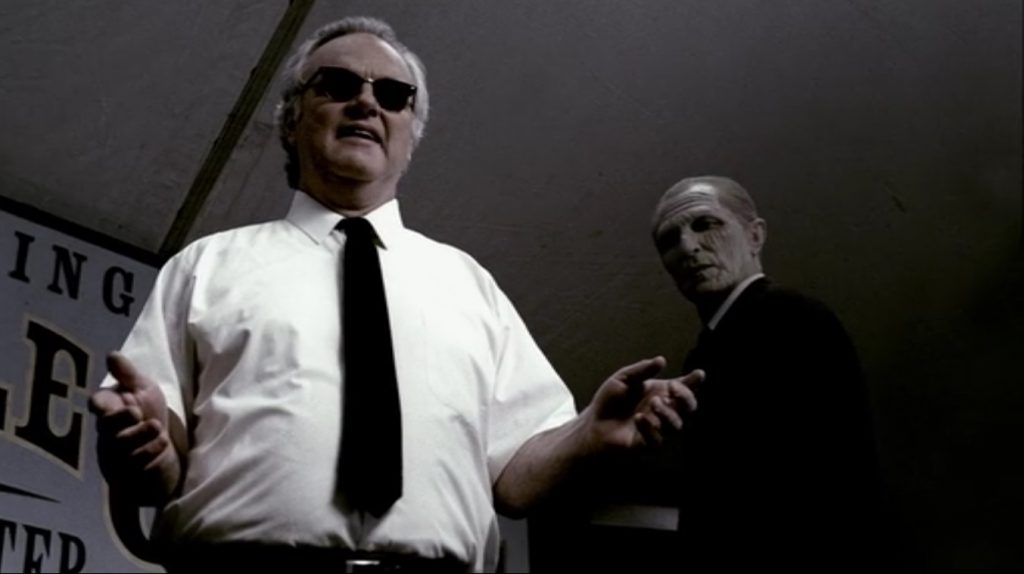 Image shows Pastor Le Grange, a portly man with gray hair and dark glasses. He's wearing a white short-sleeved dress shirt with a black tie. He's holding his hands outstretched. Behind him, a creepy dude in a black suit with a very wrinkled and deathly face is staring down at the floor, where the camera is aimed from. His expression is that of an English butler who has just witnessed someone do something of a lower class very uncouth.