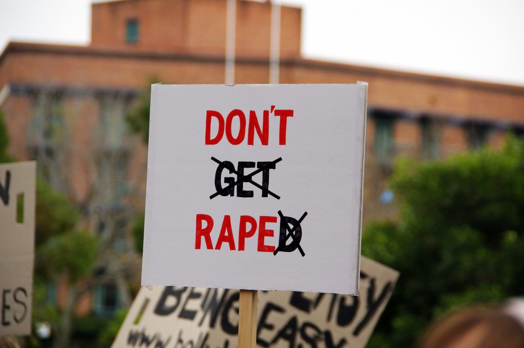 Image shows a protest sign. The letters spelled out Don't Get Raped. Get and D are in black and have been crossed out. Don't Rape is in red.