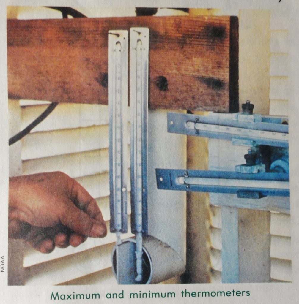 Image shows a double set of maximum and minimum mercury thermometers nailed to a wooden plank inside of a white slatted enclosure. There's a hand reaching toward the first set of thermometers. It all looks tres last century.