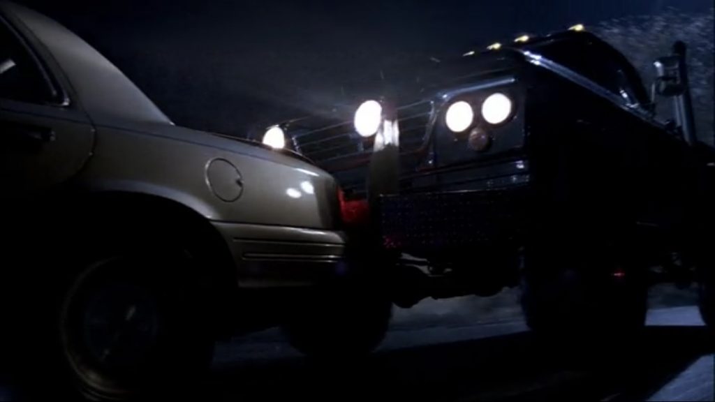 Screen capture shows a car chase by night. A monster truck with a huge rack of headlights is tailgating and about to ram a tan sedan.