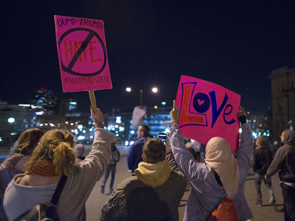 Image shows women holding up signs at a protest. We are viweing them from behind. One woman has a blond ponytail and a pink sign saying Dump Trump. In the center, the word Hate is written in red and has a circle with a line through it. Below is the hastag #notmypresident. Another person stands in the middle, not holding a sign. Beside them is a woman in hijab holding up a hot pink sign with the word LOVE written on it in huge multi-colored letters. More protestors and the city can be seen beyond.