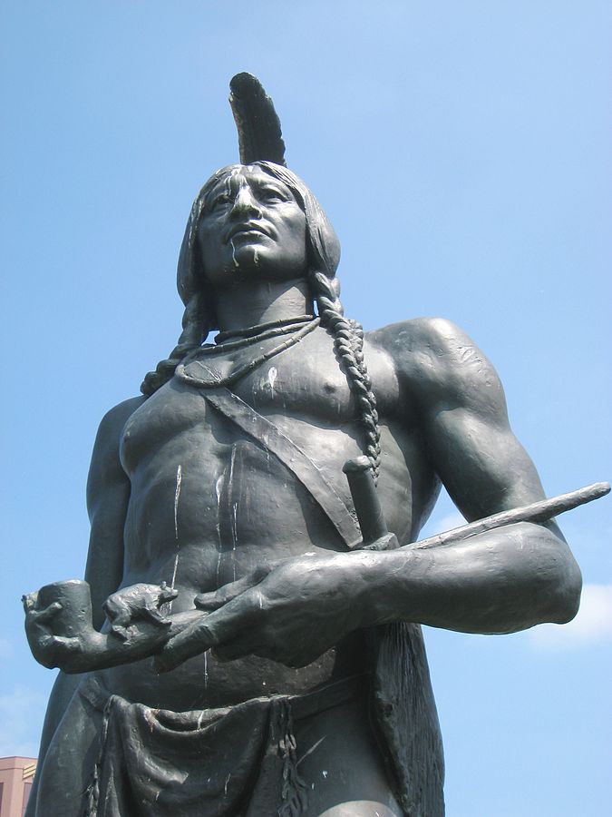 Image shows a bronze statue of a man with two long braids and a single feather inhis hair. He's holding a peace pipe.