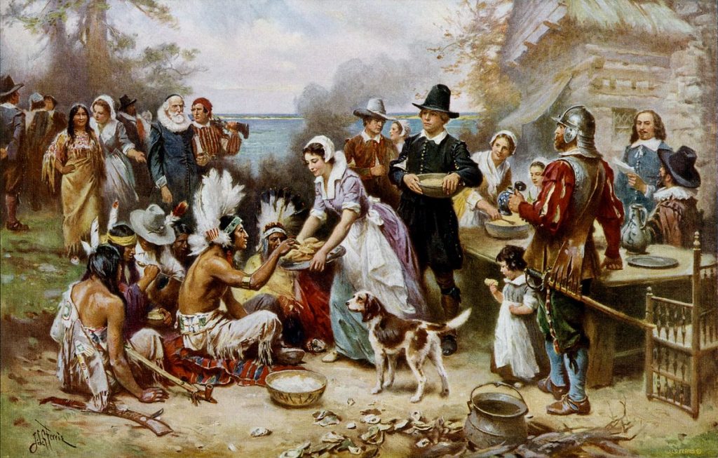 Image is a colorful painting showing Pilgrims and Wampanoag eating together. The Wampanaog are sitting on the ground, wearing feathered headdresses. One is being offered a plate of bread by a Pilgrim woman while a dog and child look on. The sea is visible in the background.