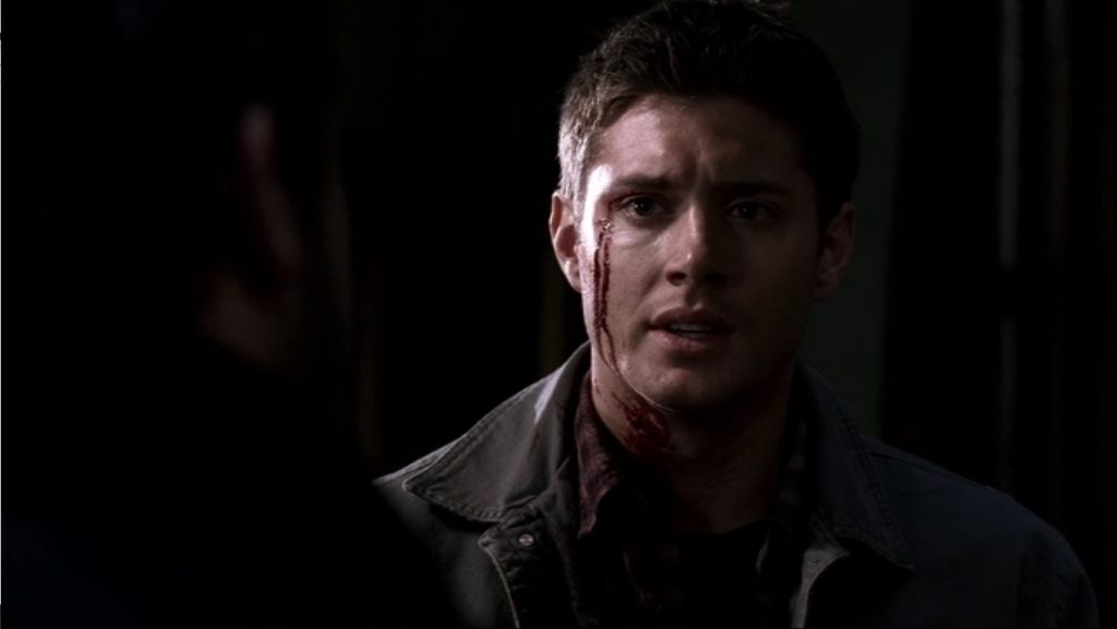 Screenshot shows Dean looking at his dad. He's got blood streaked down the left side of his face. Everything behind him is in darkness. He's looking toward his dad with a slightly furrowed brow and his mouth is slightly open, a look that reeks of apologetic fear.
