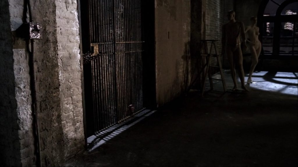 Screenshot shows an abandoned warehouse-type building interior. There's an elevator shaft with a grate over it to the left. Further along the wall with the shaft, there's a stepladder. Beside the ladder is a pair of headless, naked mannequins. One, which has a masculine body type, is posed as if it's taking a step to the side, arm out casually. The feminine mannequin is striking a sexy pose with its hip thrust to the side, knee bent, and one hand placed on its opposite hip.