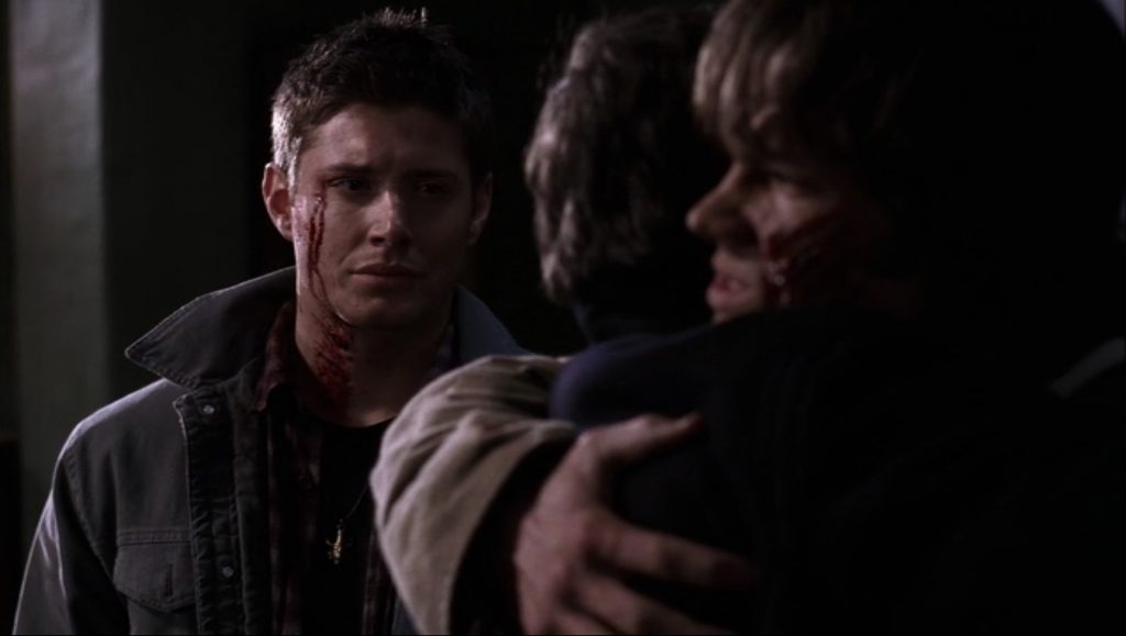 Screenshot shows Sam hugging John, while Dean looks on with his lips pursed as if he's about to cry. Blood trickling from the corner of his eye makes it look like he's been crying blood.