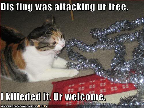 Image shows a calico cat biting a string of silver tinsel on the floor. Caption says, "Dis fing was attacking ur tree. I killeded it. Ur welcome."