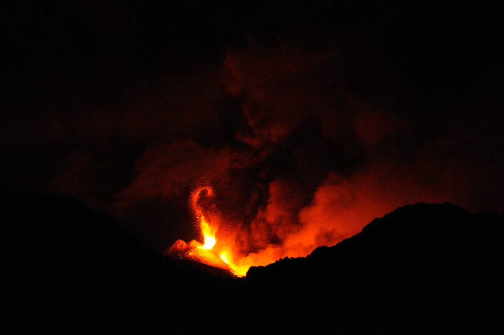 Image shows the black, jagged silhouette of the crater. Inside, clouds of steam and ash rise, lit by orange lava in the crater. One bit of the cloud on the center left is brighter than the rest, and is curled over like a tentacle.