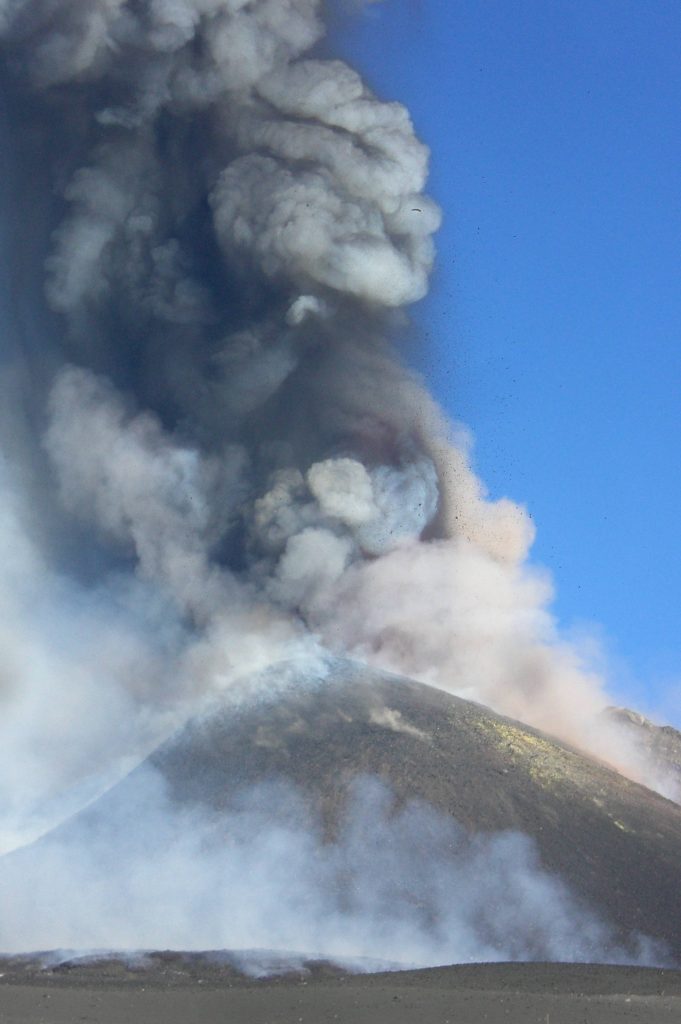 Image shows the summit of Etna with a tall, narrow, gauzy gray eruption column rising from it against the blue sky.