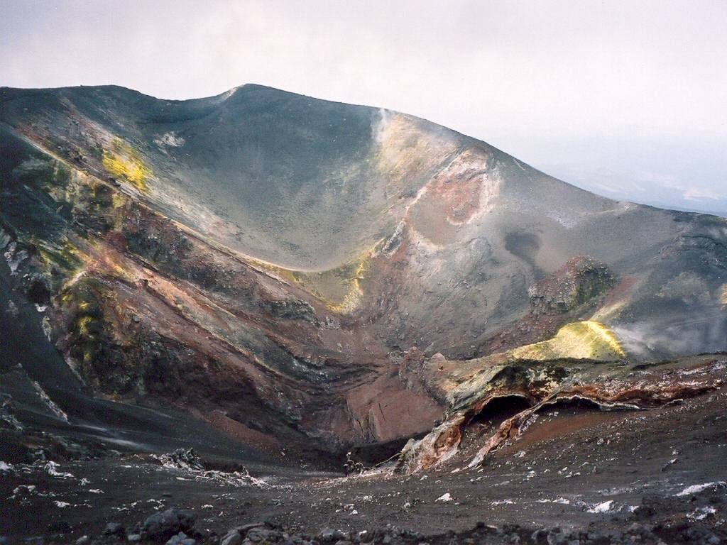Image shows a crater of Mount Etna. In the foreground there is an ash-covered dip, with the summit rising behind it. The scene is in various shades of gray, brown, and black volcanic materials, with splashes of yellow sulfur.