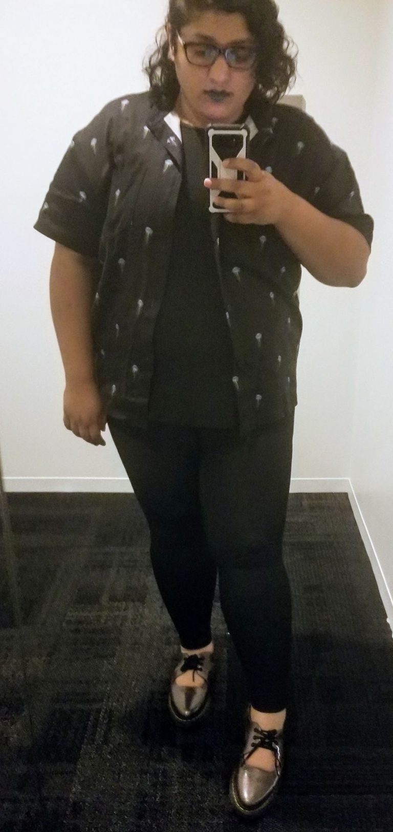 a medium brown skinned person with dark curly hair wearing an open button-up shirt with a black shirt under, dark denim leggings, and laced pointy pewter shoes