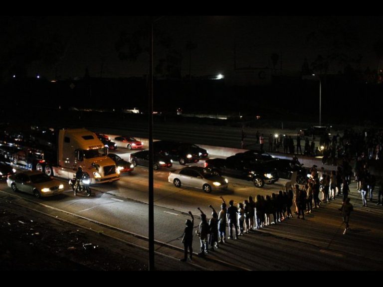 A line of people block the traffic on a freeway from moving. It's evening, so they are lit up by headlights.