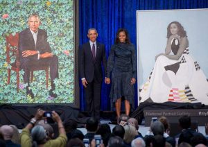 Image of former President Barack Obama and his wife Michelle Obama taking center stage at the unveiling of their official portraits. Barack Obama on the left, is flanked by his photo. Michelle Obama, holding her husbands hand, is flanked by her portrait. In front of them stand dozens of reporters.