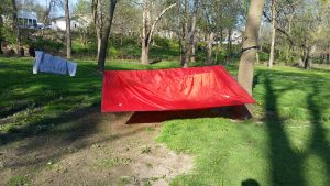 Image is of a red tarp over a hammock (not seen). The tarp is over a cord run between the trunks of two trees. More trees are visible in the background.