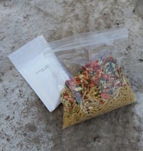 Image shows dry noodles and vegetables in a ziplock baggie, with an envelope of cheese powder next to it.