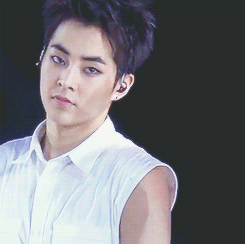 A Korean man, Xiumin from the group EXO, in a white sleeveless shirt on a black backgroud, rolls his head up and faces the right.