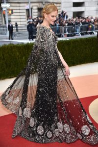 A white woman, Nicole Kidman, poses from the back in a sparkly black dress with a cape with moons at the bottom.