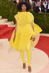 A black woman, Solange Knowles, poses in a yellow dress that resembles several crinkled fabric folded over and over.