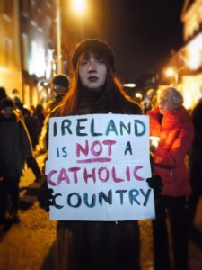 Picture of a woman standing on a street in the dark. She's wearing a coat, hat and gloves. She's holding a sign saying "Ireland is NOT a Catholic country"
