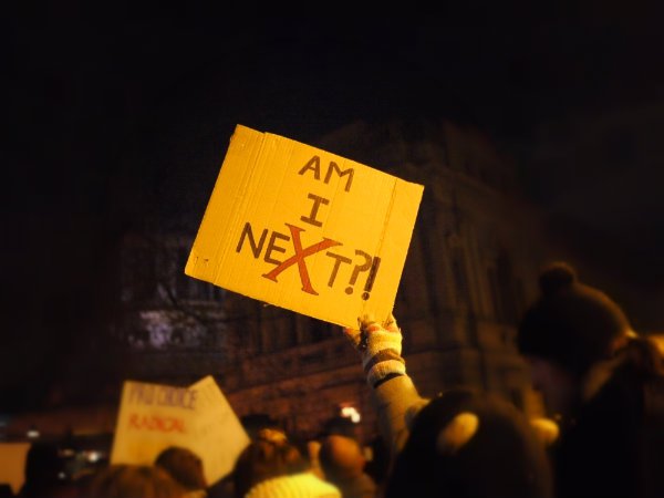 A gloved hand holds up a sign saying "Am I NeXt?"