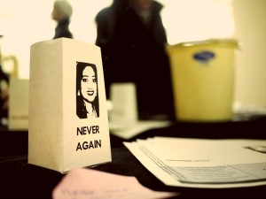 A picture of a paper tealight-holder on a table with some papers. Printed on the holder is a young woman's face (Savita Halappanavar), and the words "Never Again"