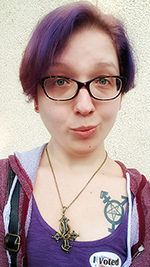 Photo of a woman standing, with short purple hair, glasses, a purple shirt and maroon hoodie, a pendant of an inverted cross.